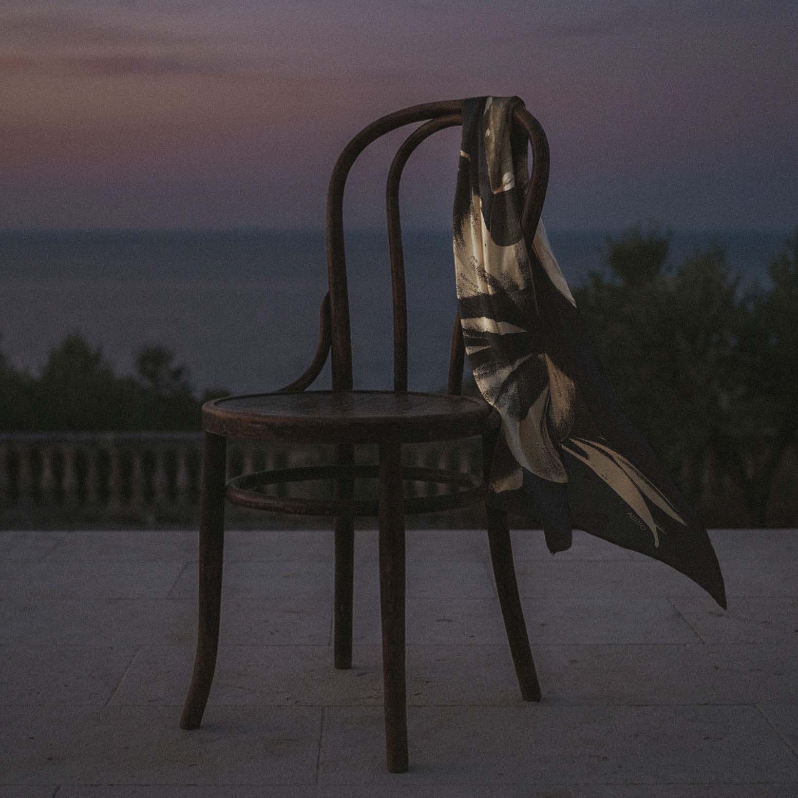 Foulard en soie Nuitée 90 Alietto drapé sur une chaise en terrasse face à la mer